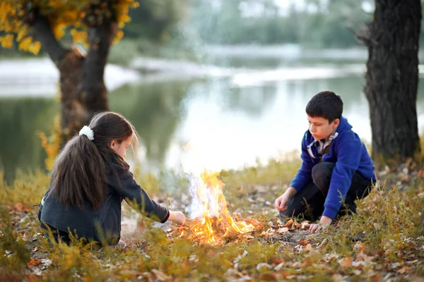 boy and girl sitting on the river Bank, make a fire, autumn forest at sunset, beautiful nature and reflection of trees in the water