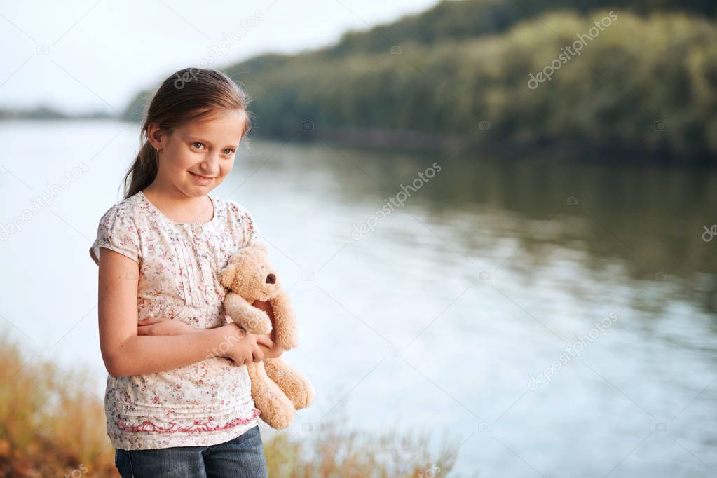 the girl child with toy bear walking along the path in the forest at sunset, beautiful river and landscape