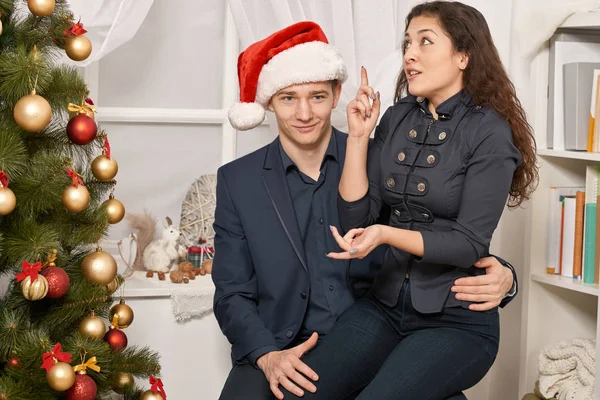 Romantic couple having fun - the girl is sitting on Santa, wants many gifts and makes wishes. Christmas tree with holiday decoration, new year theme
