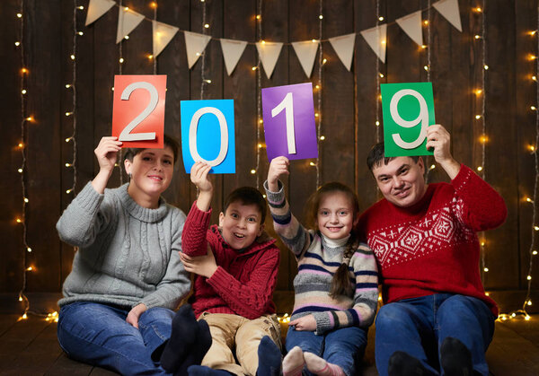 family posing with 2019 new year text, sitting on dark wooden background with christmas lights and flags, and having fun. Winter holiday concept.