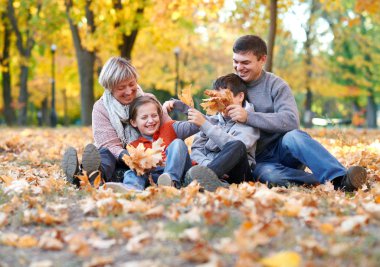 Sonbahar Şehir Parkı düşen yapraklar üzerinde mutlu aile sit. Çocuklar ve ebeveynler poz, gülümseyen, oyun ve eğlenmek. Parlak sarı ağaçlar.