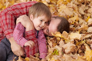 Baba ve oğul sarı yapraklarda yalan ve sonbahar şehir parkında eğleniyor. Poz, gülüyor, oynuyor. Parlak sarı ağaçlar.