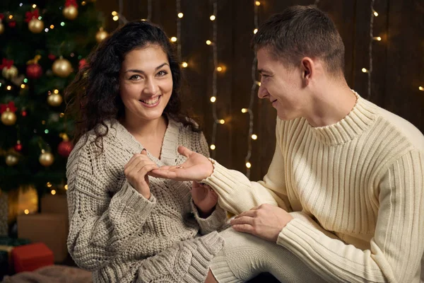 男子给女孩一个订婚戒指 夫妻是在圣诞节的灯光和装饰 穿着白色 冷杉树在黑暗的木制背景 寒假的概念 — 图库照片