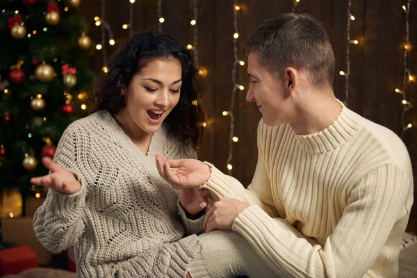 男子给女孩一个订婚戒指 夫妻是在圣诞节的灯光和装饰 穿着白色 冷杉树在黑暗的木制背景 寒假的概念 — 图库照片