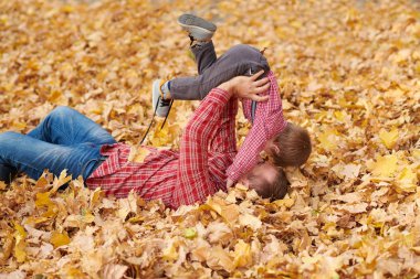 Baba ve oğul sarı yapraklarda yalan ve sonbahar şehir parkında eğleniyor. Poz, gülüyor, oynuyor. Parlak sarı ağaçlar.