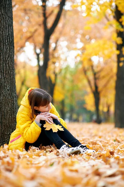 秋の公園で木の近くに悲しい十代の少女が座っています 明るい黄色の葉や木 — ストック写真