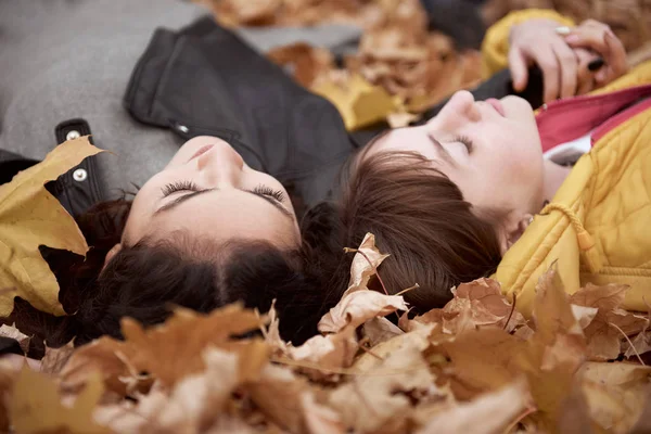 Pretty woman and teen girl are posing in autumn park. They are lying on fallen leaves. Beautiful landscape at fall season.