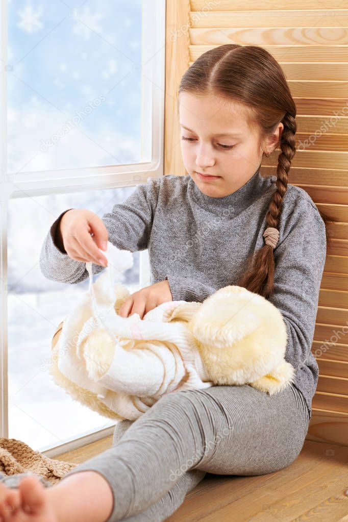 Child girl is sitting on a window sill and playing with bear toy. Beautiful view outside the window - sunny day in winter and snow.