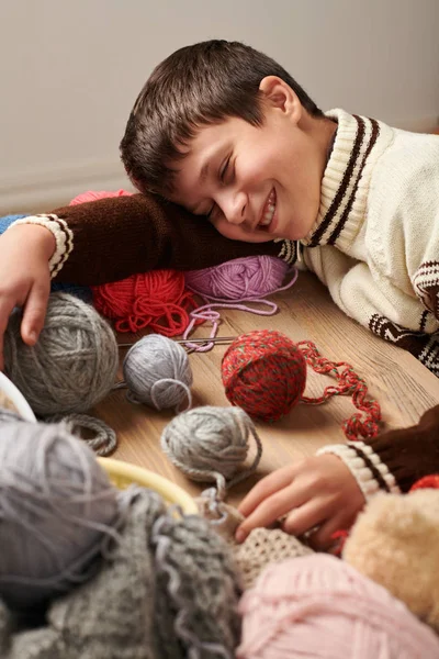 Child boy is learning to knit. He is dreaming and closing eyes. Colorful wool yarns are on the wooden table.