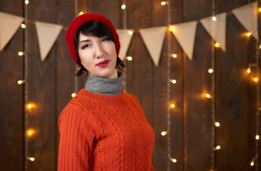 Güzel kadın koyu ahşap arka plan, Noel ışıkları ve bayraklar, tatil kavramı üzerinde poz. Kırmızı örgü şapka ve kazak giymiş.