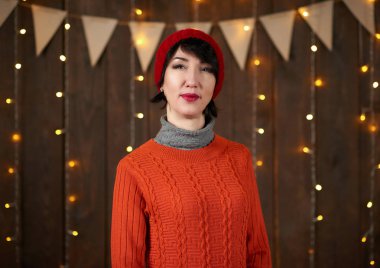 Güzel kadın koyu ahşap arka plan, Noel ışıkları ve bayraklar, tatil kavramı üzerinde poz. Kırmızı örgü şapka ve kazak giymiş.