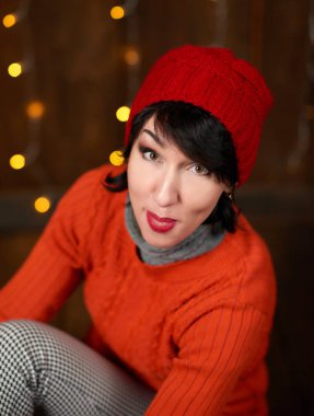 Güzel kadın koyu ahşap arka plan, Noel ışıkları ve bayraklar, tatil kavramı üzerinde poz. Kırmızı örgü şapka ve kazak giymiş.