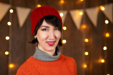 Güzel kadın koyu ahşap arka plan, Noel ışıkları ve bayraklar, tatil kavramı üzerinde poz. Kırmızı örgü şapka ve kazak giymiş.