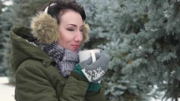 女人在冬天的森林里喝热茶或咖啡 她头上戴着皮草耳罩 美丽的风景与雪的冷杉树 — 图库视频影像