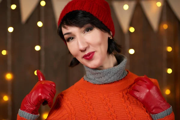 Güzel kadın koyu ahşap arka plan, Noel ışıkları ve bayraklar, tatil kavramı üzerinde poz. Kırmızı örgü şapka ve kazak giymiş.
