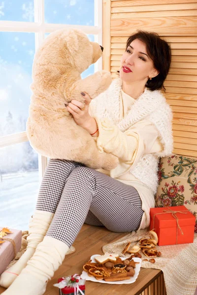 Woman is sitting with bear toy on a window sill. Beautiful view outside the window - sunny day in winter forest and snow.