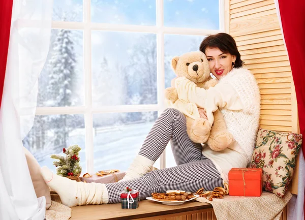 Woman is sitting with bear toy on a window sill. Beautiful view outside the window - sunny day in winter forest and snow.