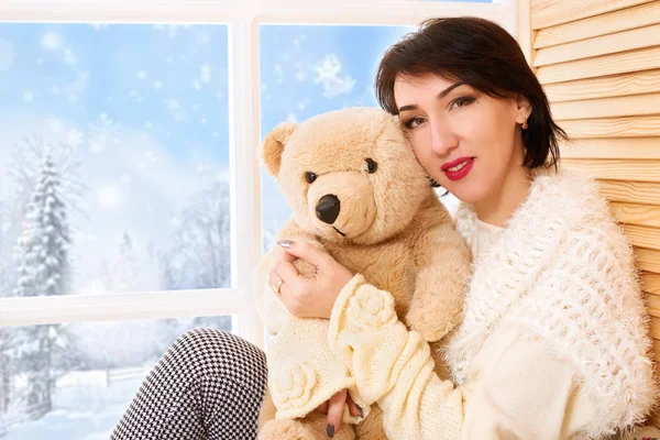 Woman is sitting with bear toy on a window sill. Beautiful view outside the window - sunny day in winter forest and snow.