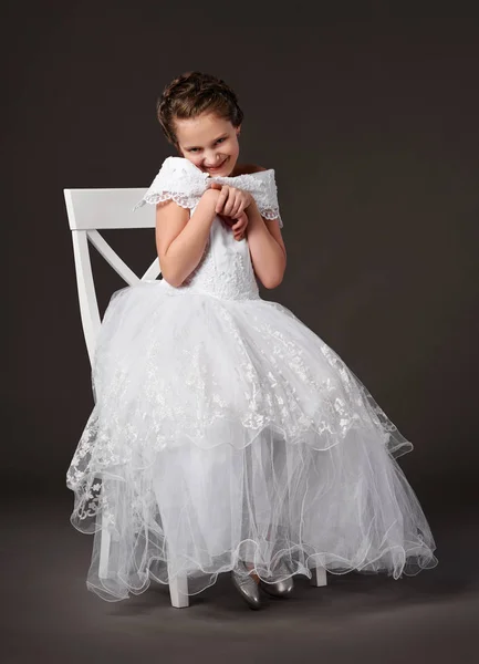 Маленькая девочка одета в белое бальное платье и сидит на стуле, темный фон — стоковое фото