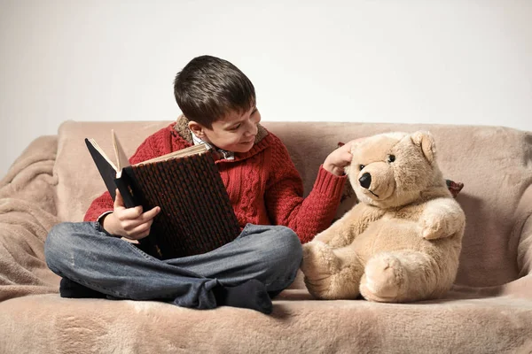 boy reads a book, sits on the sofa with a bear toy