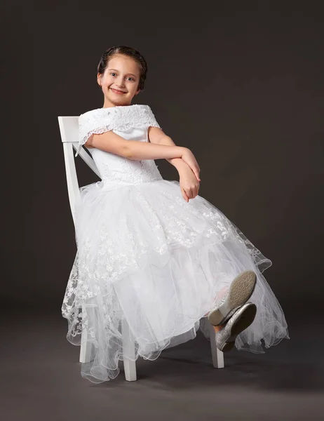 Niña vestida con un vestido de bola blanco y sentada en la silla, fondo oscuro — Foto de Stock