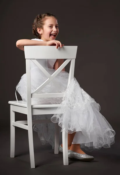 Liten flicka klädd i en vit balklänning och sitter på stolen, mörk bakgrund — Stockfoto