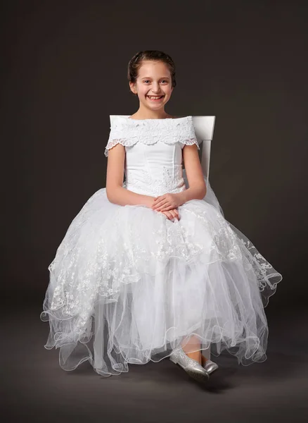Liten flicka klädd i en vit balklänning och sitter på stolen, mörk bakgrund — Stockfoto