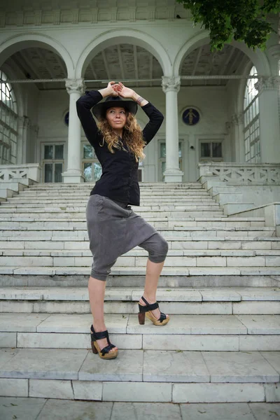 Chica de moda vestida con camisa negra, sombrero y pantalones anchos posando cerca de la vieja casa blanca —  Fotos de Stock