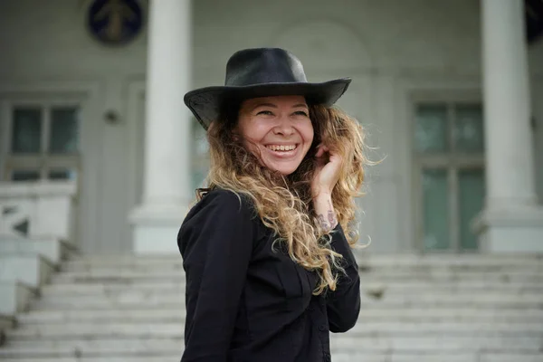 fashionable girl dressed in black shirt, hat and wide trousers posing near old white house