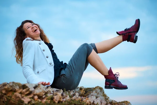 Chica de moda vestida con chaqueta blanca y pantalones anchos posando al atardecer fondo del cielo, se sienta en la piedra — Foto de Stock