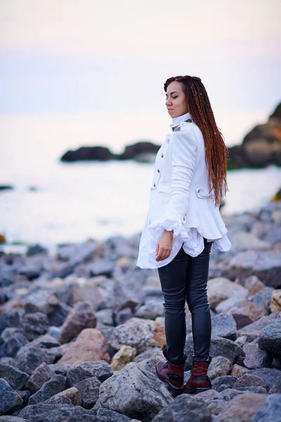 Dreadlocks chica de moda vestida con chaqueta blanca y pantalones de cuero negro posando cerca del mar por la noche —  Fotos de Stock