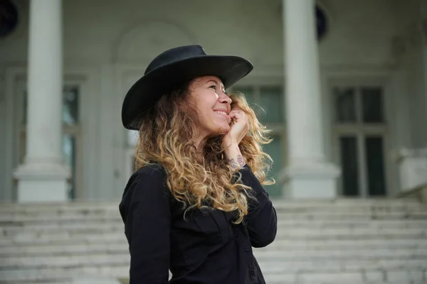Modna dziewczyna ubrana w czarną koszulę, kapelusz i szerokie spodnie pozowanie w pobliżu Starego białego domu — Zdjęcie stockowe