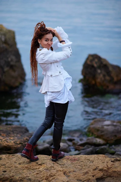 Dreadlocks chica de moda vestida con chaqueta blanca y pantalones de cuero negro posando cerca del mar por la noche —  Fotos de Stock