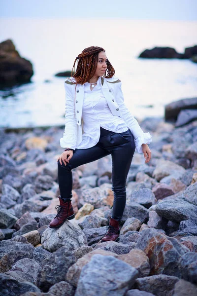 Ντρήλοκ μόδας κορίτσι ντυμένος με λευκό μπουφάν και μαύρο δερμάτινο παντελόνι που ποζάρει κοντά στη θάλασσα το βράδυ — Φωτογραφία Αρχείου