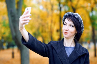 Akıllı telefonla selfie çeken ve sonbahar şehir parkında eğlenen güzel kadın, sonbahar sezonu, sarı yapraklar