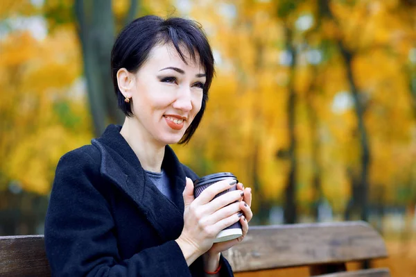 Mulher bonita sentada com café no parque da cidade de outono, estação de outono, folhas amarelas — Fotografia de Stock