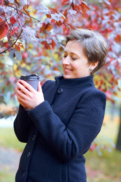 Krásná žena s papírovým šálkem čaje nebo kávy v podzimním městském parku, podzimní sezóna — Stock fotografie