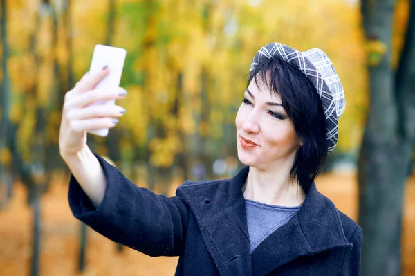 Красива жінка бере селфі за допомогою смартфона і розважається в осінньому міському парку, осінній сезон, жовте листя — стокове фото