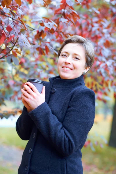 Bella donna in posa con tazza di carta di tè o caffè nel parco della città di autunno, stagione autunnale — Foto Stock