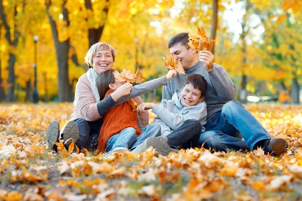 Glückliche Familie, die auf fallendem Laub sitzt, spielt und sich im herbstlichen Stadtpark vergnügt. Kinder und Eltern haben gemeinsam einen schönen Tag. helles Sonnenlicht und gelbe Blätter an Bäumen, Herbstzeit. — Stockfoto