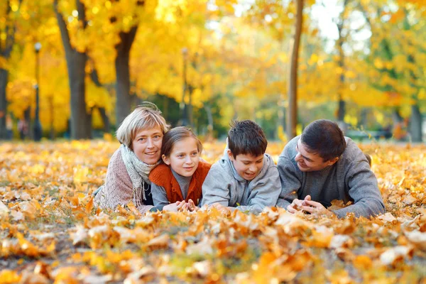 Família feliz deitada em folhas caídas, brincando e se divertindo no parque da cidade de outono. Crianças e pais juntos tendo um bom dia. Luz solar brilhante e folhas amarelas em árvores, estação de outono . — Fotografia de Stock