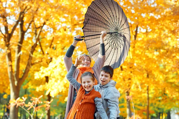 Familia feliz posando bajo el paraguas, jugando y divirtiéndose en el parque de otoño de la ciudad. Los niños y los padres juntos tienen un buen día. Luz del sol brillante y hojas amarillas en los árboles, temporada de otoño . — Foto de Stock