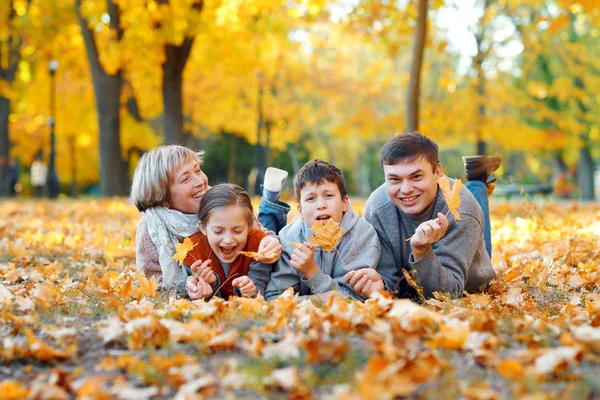 Familia feliz acostada sobre hojas caídas, jugando y divirtiéndose en el parque de otoño de la ciudad. Los niños y los padres juntos tienen un buen día. Luz del sol brillante y hojas amarillas en los árboles, temporada de otoño . —  Fotos de Stock