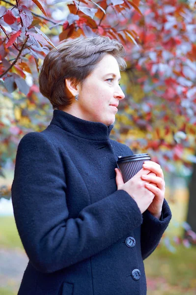 Beautiful woman posing with paper cup of tea or coffee in autumn city park, fall season — Stock Photo, Image