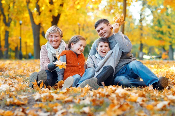 Šťastná rodina sedí na padlých listech, hrají a baví se v podzimním městském parku. Děti a rodiče spolu mají hezký den. Světlé sluneční světlo a žluté listy na stromech, období pádu. — Stock fotografie