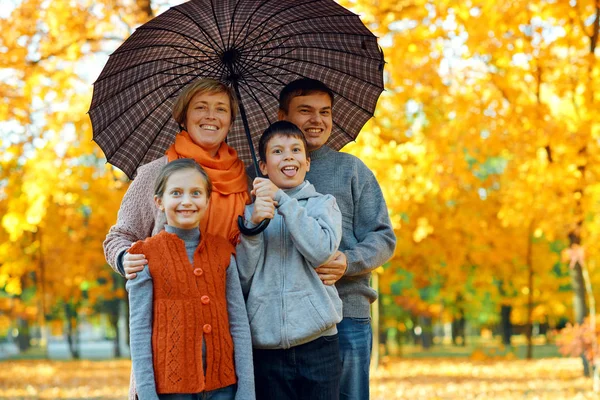 Happy family posing under umbrella, playing and having fun in autumn city park. Children and parents together having a nice day. Bright sunlight and yellow leaves on trees, fall season.