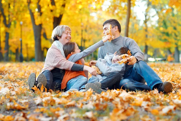 Szczęśliwa rodzina siedzi na opadłych liściach, grając i bawiąc się w jesiennym parku miejskim. Dzieci i rodzice razem o miłym dniu. Jasne światło słoneczne i żółte liście na drzewach, jesień sezonu. — Zdjęcie stockowe