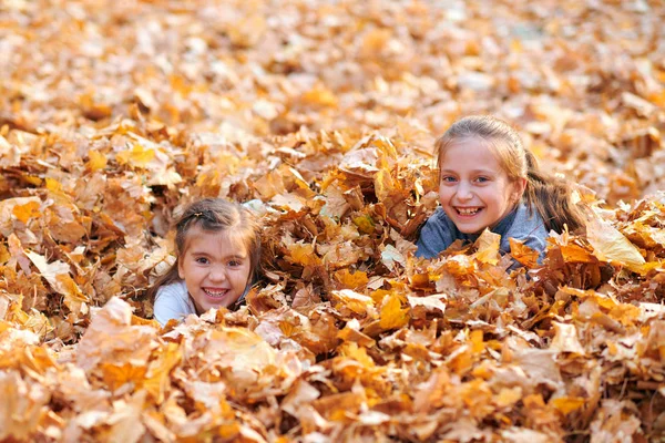 Lyckliga barn som leker i högen med gula löv, poserar, ler och har roligt i höst stadsparken. Ljust gula träd och löv — Stockfoto