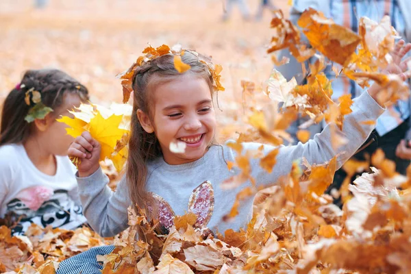 Szczęśliwe dzieci bawią się w stos żółtych liści, stwarzając, uśmiechając się i zabawy w jesiennym parku miejskim. Jasne żółte drzewa i liście — Zdjęcie stockowe