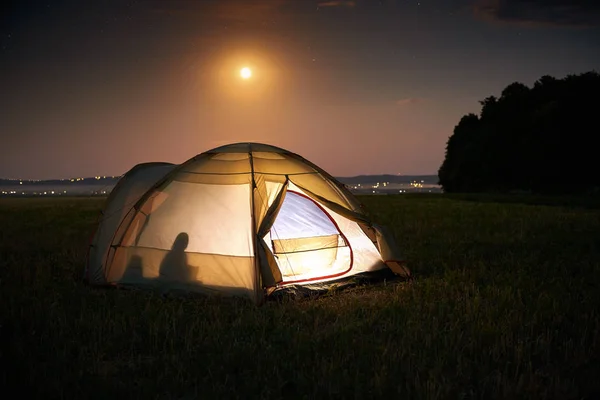 Concetto di viaggio e campeggio - tenda da campeggio di notte sotto un cielo pieno di stelle. Tenda arancione illuminata con una persona all'interno. Bella natura - campo, foresta, pianura. Luna e chiaro di luna — Foto Stock